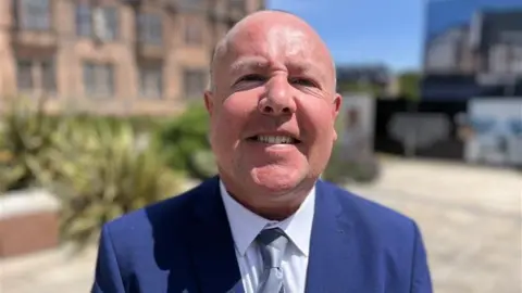 A man stood outside office buildings wearing a blue suit, white shirt and silver tie. He is bald and is smiling at the camera. The background is blurred, but you can see some bushes and plants.