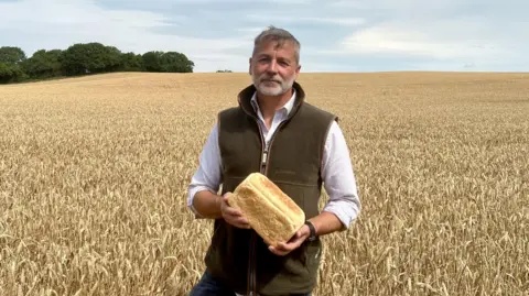 A photo of a man standing in a field with a loaf of bread, he is looking directly down into the camera. He is wearing a shirt and a vest jacket. 