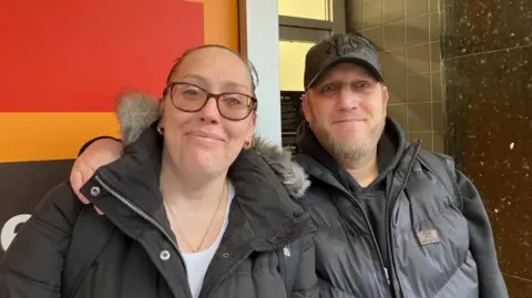 Vikki Irwin/BBC A woman and a man are standing next to each other. The man has his hand over the woman's shoulder. The woman is wearing a puffed jacket with a fur lined hood and is black. She is wearing glasses has gold earrings and necklace visible. The man is wearing a black cap also has a puffed jacket in black. He is also smiling at the camera. They are standing on the street and there is a red and orange background. 