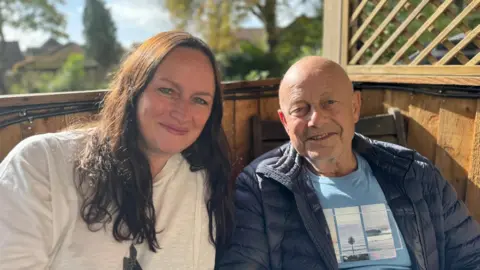 A man and a woman smiling at the camera while sitting underneath a wooden shelter in a garden.