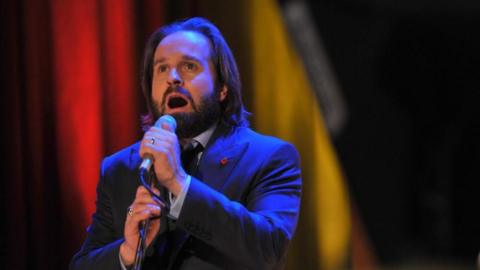 Alfie Boe performing, wearing a midnight blue suit, with a white shirt and poppy pin badge 