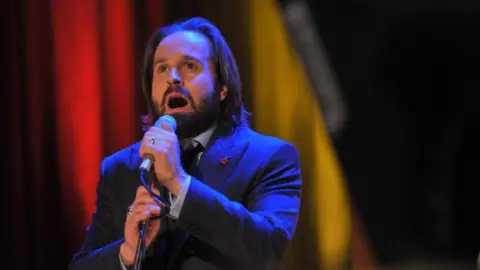 BBC Alfie Boe performing, wearing a midnight blue suit, with a white shirt and poppy pin badge 