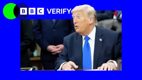US President Donald Trump wearing a black suit with a blue tie sat in the Oval Office with a pen in his hand.