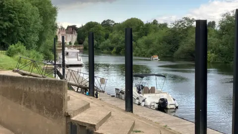 The River Ouse at Bishopthorpe with Bishopthorpe Palace in the background. There are three boats on the river, two of them are moored. There are trees on either side of the river.