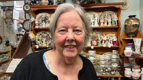 A woman with tied-back grey hair smiles at the camera. She is wearing a black cardigan and standing in front of shelves covered with cups, plates and ornaments.