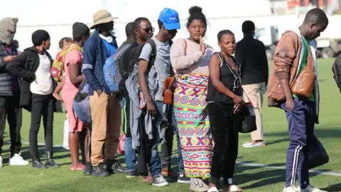People standing in a queue on some grass. Some are carrying backs on their backs and wearing hats.