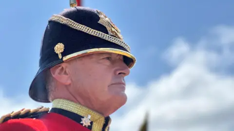 Close-up of a trumpeter, who is wearing a red tunic with gold trim. His helmet is black with a gold spike and decoration. There is blue sky with some cloud behind him.