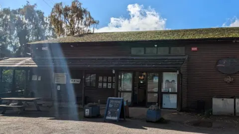 The image is of the visitor centre before it was burgled. It shows a brown building with glass doors at the left of the image. The sky is blue with a few white clouds. 