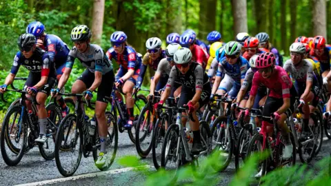 A tightly packed group of road cyclists on a road surrounded by woodland.
