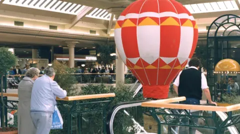 Metrocentre The upper balcony of the shopping centre with people leaning over the wooden bannister. In the open space in the centre there is a small inflatable red, white and yellow hot air balloon. 