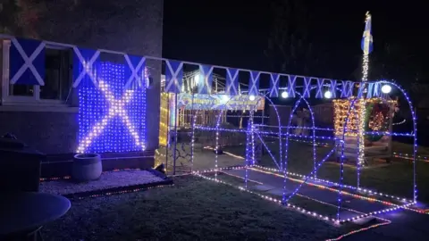 Sandra Lee Myles Saltire flag and blue light decorations for St Andrew's Day. There is bunting, a flag on the side of the house and a blue light tunnel.