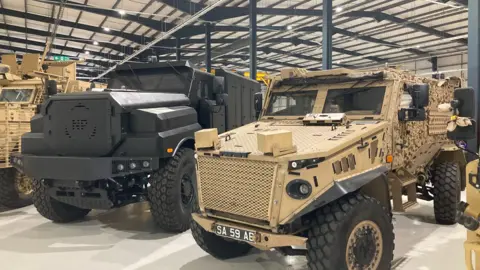 A black Foxhound vehicle and a beige Ridgeback vehicle sit site by side in the factory. The factory has a metal, ridged roof which is grey overhead.