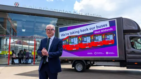 West Midlands Combined Authority West Midlands mayor Richard Parker, standing in front of a digital ad van with the slogan 'We're taking back our buses'. Wolverhampton bus station can be seen in the background and a group of young people wearing rucksacks are standing near the entrance.