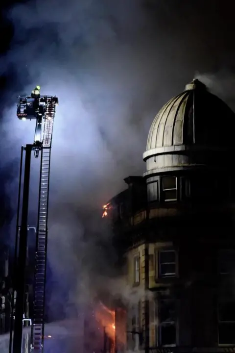 Michelle Cohen A firefighter on an aerial appliance, looking over the dome. There is lots of smoke in the sky and flames can be seen at the bottom of the building. The dome is in tact.