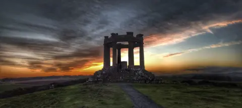 Jim Lowden Round stone war memorial made of six columns with a vivid orange, blue and grey sunset in the background.