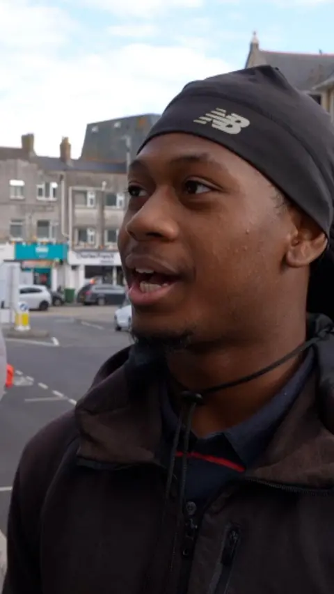 A 19-year-old man wearing a dark hat and jacket speaks to camera.