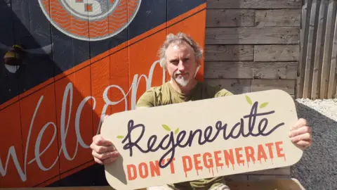 A man with wayward mid-length wavy grey hair holds up a sign that reads "Regenerate don't regenerate" as he sits outside on a gravelled area in the sunshine. The word "Welcome" is painted on a shed behind him.