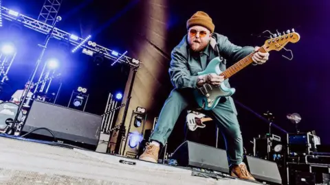 Angus Mclennan/iGigs Thom Rylance is standing on the stage, bending down towards the camera with electric guitar in his hands.
