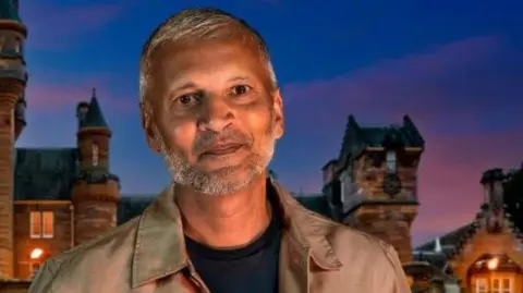 The Traitors Maz, who is wearing a black T-shirt and Kharki overshirt, is standing in front of the castle where the series of The Traitors is filmed and smiling at the camera.