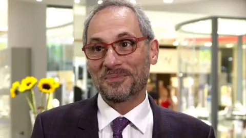 Peter Jackson, who has short grey hair and beard and red-framed glasses, stands in a shop smiling. He is wearing a dark suit and tie and a white shirt and there is a vase of sunflowers over his shoulder to the left