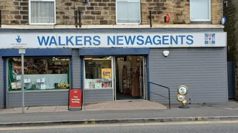 Suki Walker A traditional newsagent. Walkers Newsagents is written in large blue letters on the front of the building. Flats are above the store.