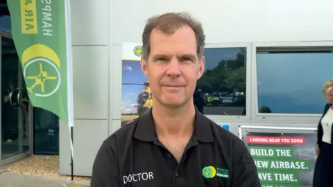 A man is looking towards the camera wearing a black polo shirt. The words 'Doctor' and 'Hampshire and Isle of Wight Air Ambulance are printed on the front. He has balding black hair.