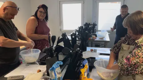 A group of people in a kitchen. The tables have cooking implements on them as well as mixing bowls which the people are stood over making socnes in them.