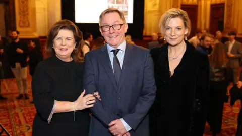 Mark Mainz Two women and a man stand in a room full of people. On the left, Elaine C Smith, who has brown hair and is wearing a black dress with jewels on the sleeves. She is linking arms with Tommy Gormley. He is smiling, wearing glasses and a blue suit. On the right is Sarah Purser, who has blonde hair which is tied back and showing off some earrings. She is wearing a black jacket and a necklace.