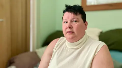 A woman sitting on a sofa wearing a white sleeveless top.
