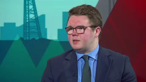 BBC Councillor Antony Mullen sits in a TV studio wearing a blue suit and green tie. He has thick wavy brown hair and wears black-rimmed glasses. The teal and red backdrop of the BBC Politics North studio can be seen behind him. 