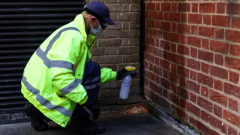 A person in a high vis jacket and wearing a face mask sprays liquid at a brick wall.