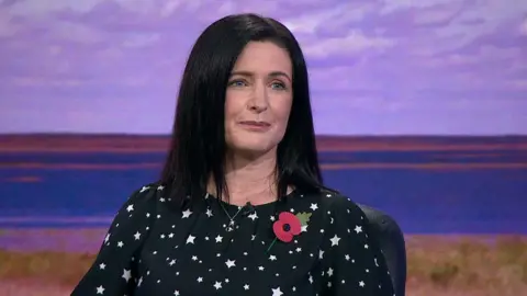 Paula Bradshaw. She has shoulder length dark hair and blue eyes, wearing a silver necklace and black top with white star print. She is wearing a poppy pin.