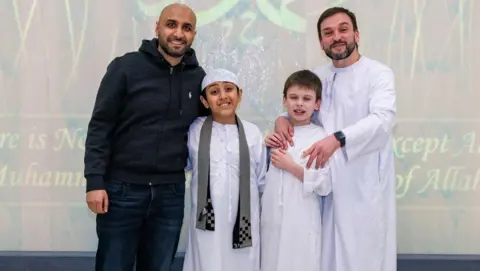 Kasim Choudhry Two men and two boys stand in front of a board. Kasim Choudhry, left, pictured with his son and Dan Harris and his son, Joshie