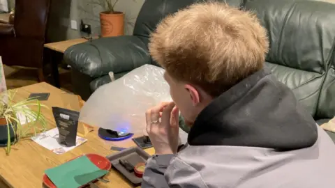 The back of a man's head, sitting at a coffee table holding a plastic bag filled with vapour to his mouth. He is wearing a light jacket with a black hood and there is a green leather sofa behind him to the right.