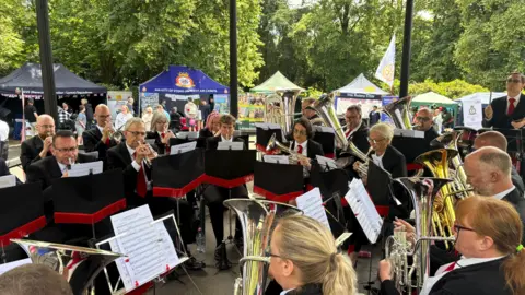 A brass band of more than a dozen people is playing a piece of music and looking at music in front of them. Trees are in the background.