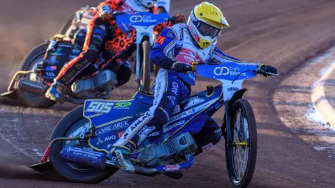 Ian Charles A speedway rider with a yellow helmet on a blue scrambler-style bike racing on a dirt track. 
