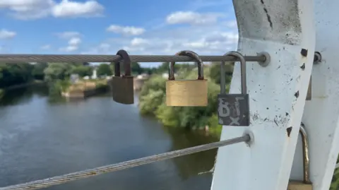 A close up of part of a dirty white and scratched steel bridge structure, with three different small padlocks hanging from the steel wires running from it