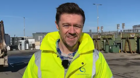 Ben Henry, wearing high-vis with the Guernsey Waste logo, stood in front of Longue Hougue Household Waste & Recycling Centre. He has short dark hair and a short beard.
