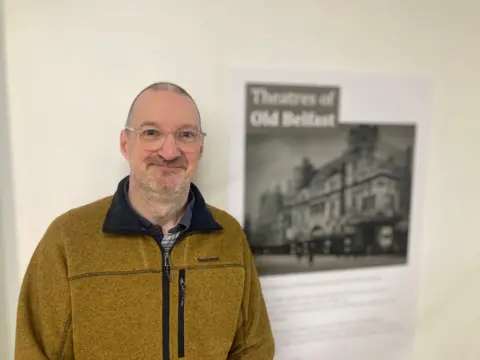 Stuart Marshall is wearing a yellow fleece and round spectacles. He is stood next to a poster which says 'Theatres of old Belfast