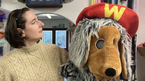 A dark-haired woman wearing a cream round-necked jumper looking at the head of a Womble costume she is holding in her left hand. Above her head is a sign saying, "stage door".