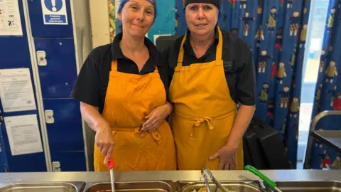 Simon Thake/BBC Two women in black hair nets and yellow aprons stand in front of metal serving dishes of food