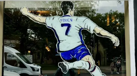 A painting of Phil Foden about to strike a ball with his right leg on the window of the Church Inn in Swinton
