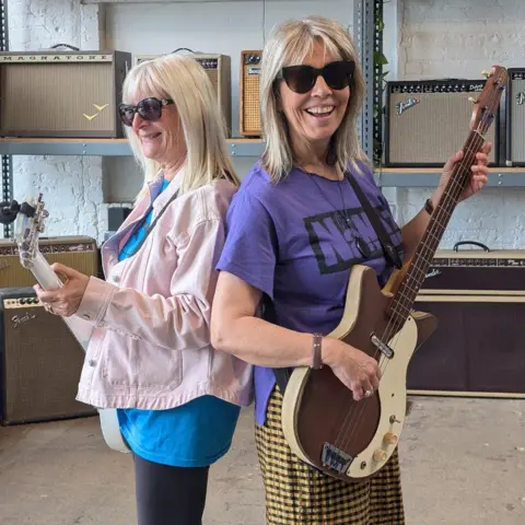 Nan Davies / Laura Robinson / BBC Two women stood back to back. One is wearing black leggings, blue t shirt and pink jacket, the other yellow and brown check trousers and a purple t shirt. They are both blonde, wearing sunglasses and holding electric guitars. Behind them are shelves with amplifiers on them.