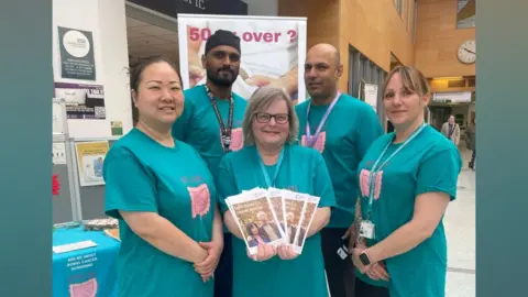North West Anglia NHS A group of five men and women stand in front of an exhibition stall about bowel cancer, all wearing matching teal-coloured T-shirts with a picture of a pink bowel on the front. They wear lanyards and a woman in the middle of the group holds up a fan of leaflets about spotting bowel cancer early. They are in the entrance to what appears to be a hospital.