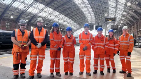 Eight people wearing orange high-vis and hard hats standing a station platform smiling at the camera