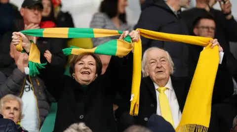 Adam Davy/PA Wire Norwich City directors Delia Smith, left, and Michael Wynn Jones cheer on their team during a Championship match at Carrow Road in April 2023. Delia is wearing a black jacket and holding a yellow and green scarf. Michael Wynn Jones is wearing a black coat and a white shirt and yellow tie. He is holding a yellow scarf. They are in front of a group of people.