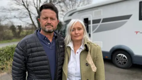 Steve Hubbard/Mr. BBC, Fenton's short hair, blue T-shirt and black Gilet stood next to Mrs. Fenton, with long hair, white hair, white shirt and stone-colored jacket. They stood next to the white RV with grey and black details.