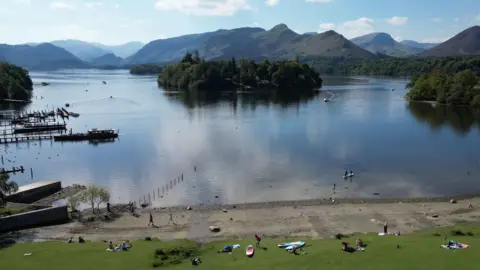 PA Media A sunny day over Derwentwater near Keswick in the Lake District. The vast lake has islands dotted around on it, which are lush with trees. There are boats speeding across the water. There are docks jutting out from the edge of the lake. And a grassy area in the foreground where people are sitting.