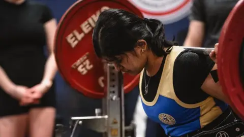 Photolab A woman balancing a barbell on her upper back with two red plates on either side, both 25kg, preparing to squat in a competition.