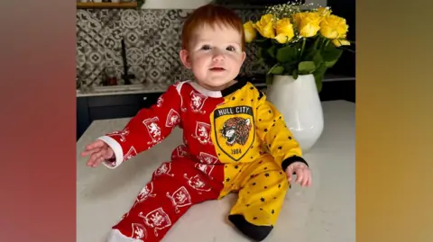 Supplied Baby Beatrice sits atop a kitchen counter wearing a half red and half yellow baby gro. Each side has the respective badges on. She has short red hair and is smiling slightly at the camera, with her hands outstretched. There is a vase of yellow flowers behind her.